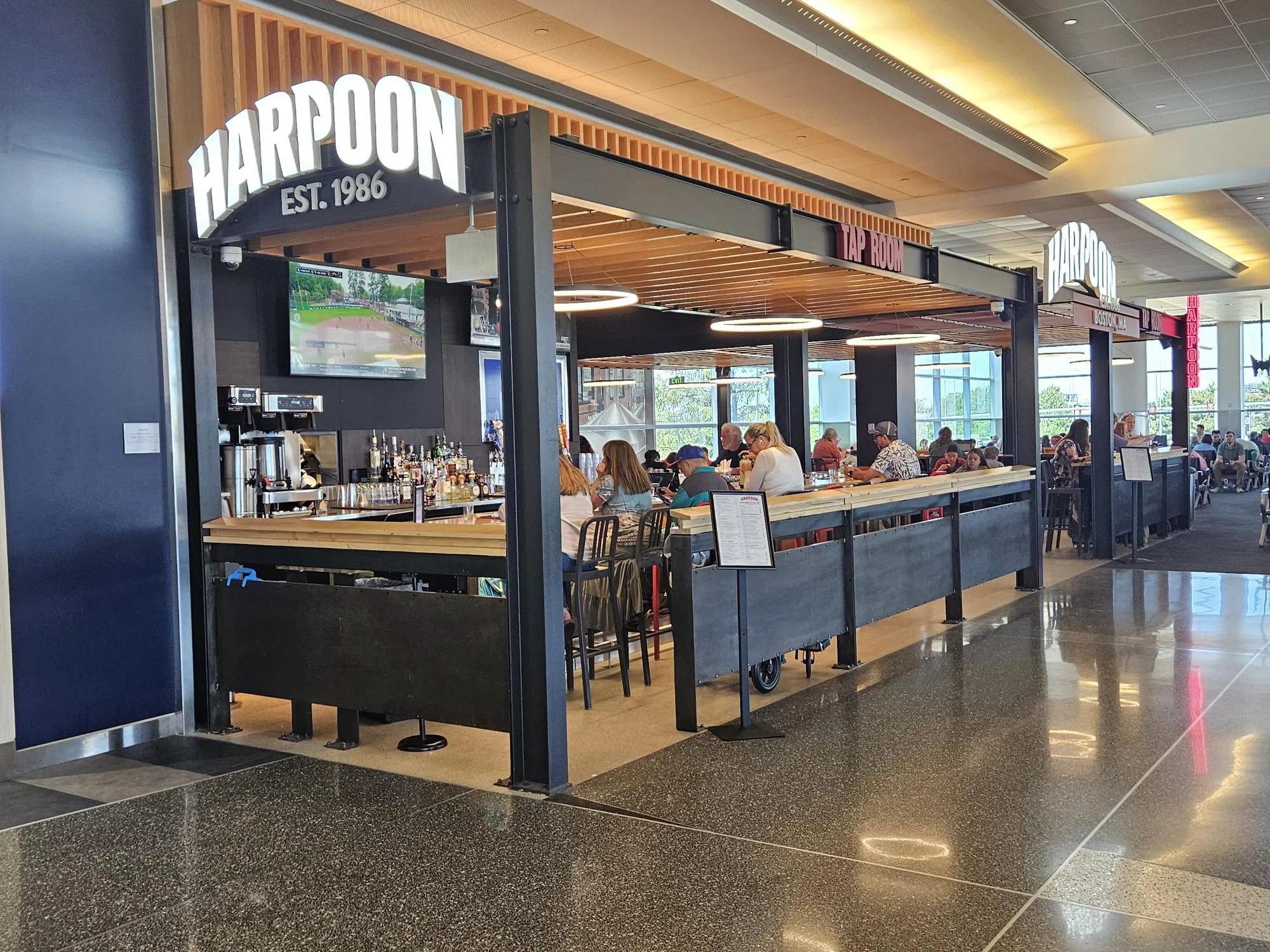 Harpoon Est. 1986 Tap Room bar in an airport concourse, with a wood slat ceiling, a black steel frame, and a wood-top bar with dark panels