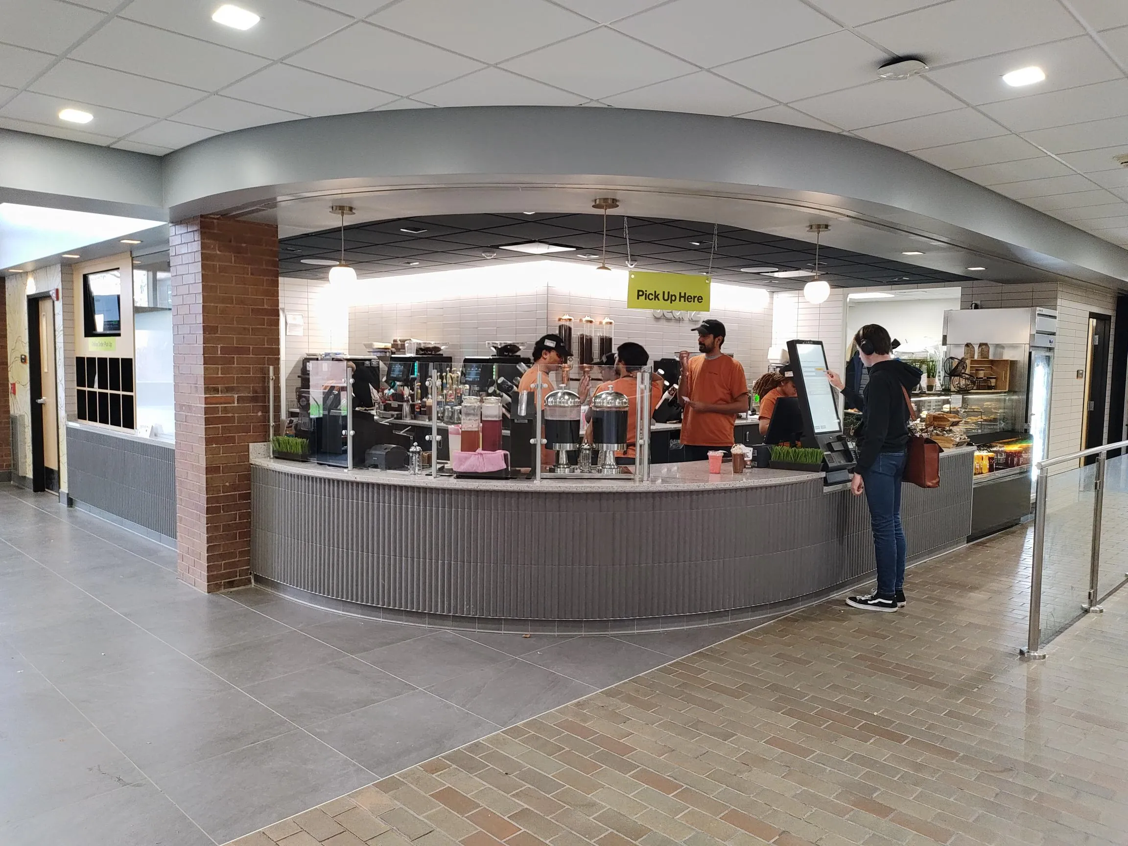 Wide view of the curved Beanz cafe service counter with grey ribbed tile, a Pick Up Here sign, and pendant lights