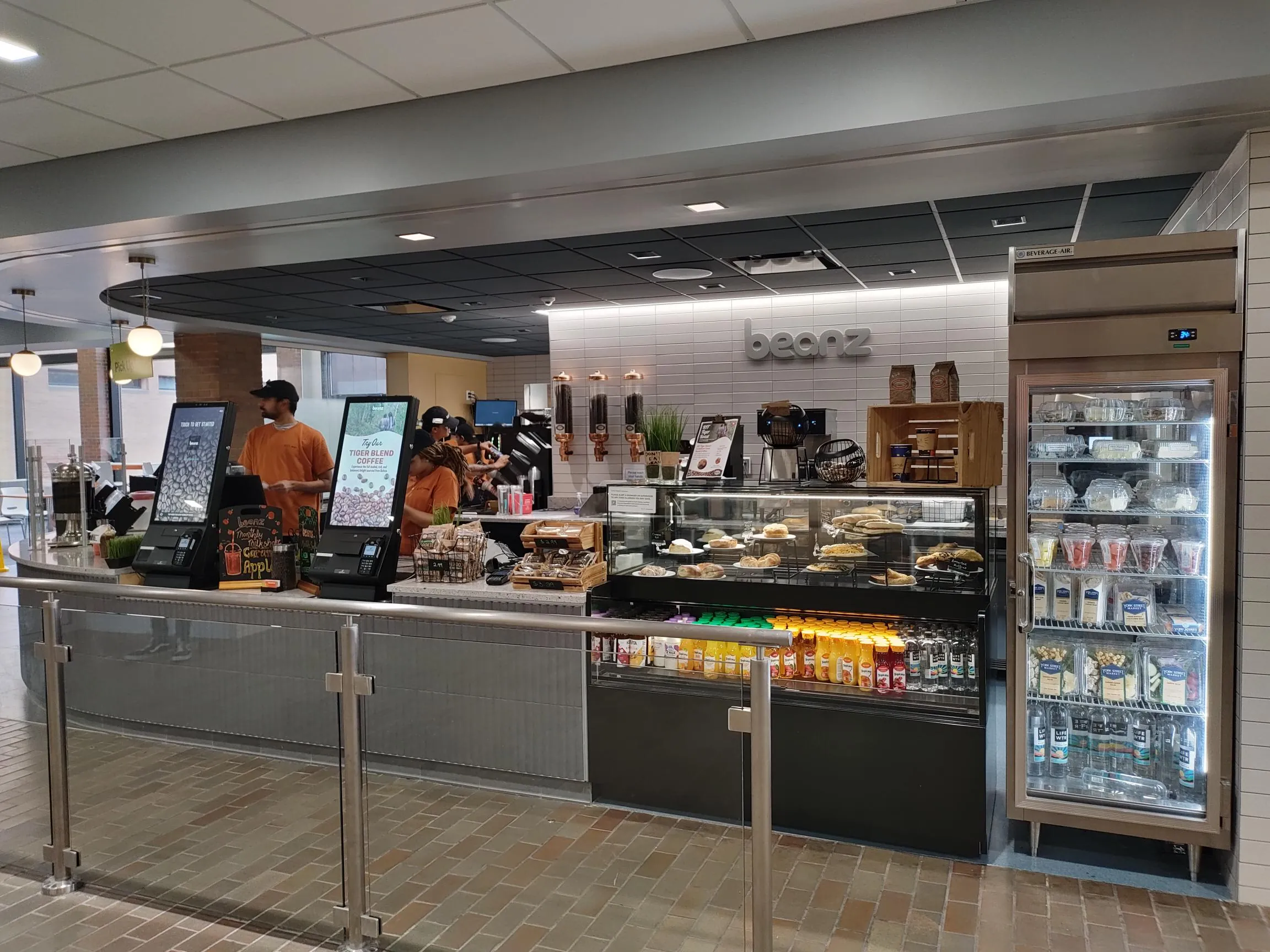 Front of the Beanz counter with ordering kiosks, a bakery display case, and a grab-and-go refrigerator